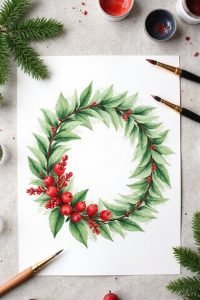 Watercolor painting of a festive wreath with green leaves and red berries on a table with paintbrushes and paints.