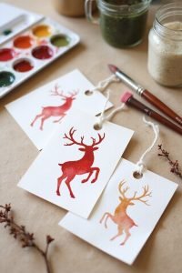 Watercolor reindeer gift tags with paint palette and brushes on a craft table.