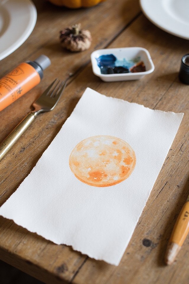 Watercolor painting of an orange moon on paper, surrounded by art supplies on a wooden table.