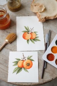 Watercolor paintings of oranges on handmade paper with art supplies on a rustic table.