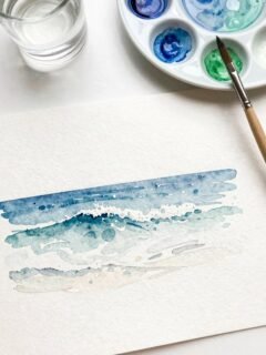 Watercolor sea painting with blues and greens, next to a paint palette, brush, and a glass of water.