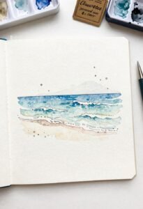 Watercolor beach painting in sketchbook with paint palette.