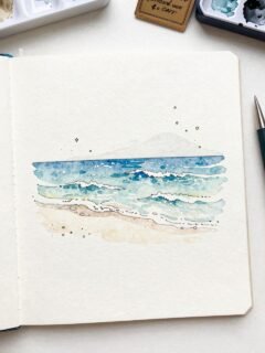 Watercolor beach painting in sketchbook with paint palette.