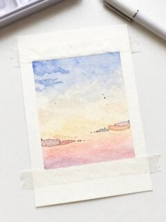 Watercolor painting of a pastel sunset with clouds taped to a white surface, next to a pen.