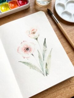 Watercolor painting of pink flowers in a sketchbook, with paint set and brush on a wooden table.